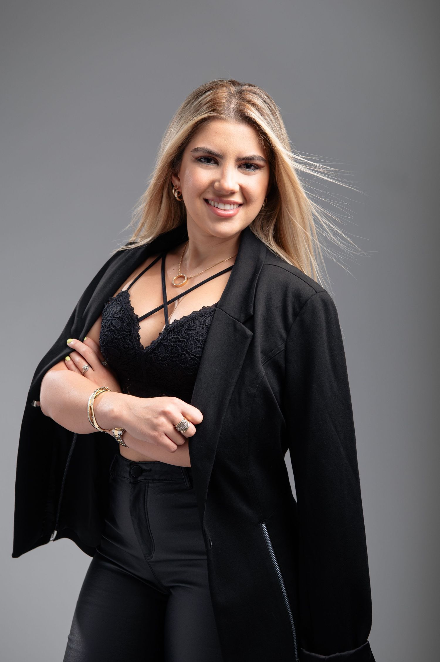 Fashion model in black outfit with flowing blonde hair against gray studio background.