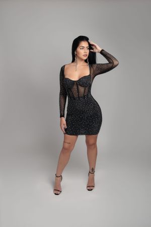 Model in fitted black sparkly dress and sheer sleeves posing against grey studio background.