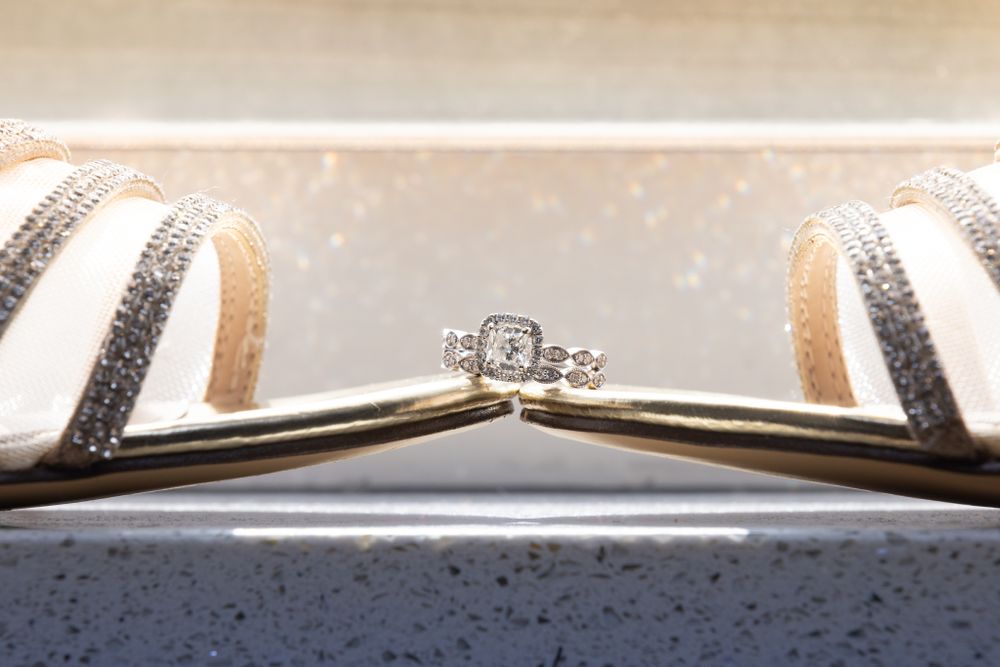 Diamond ring artistically positioned between white bridal shoes against blurred background.