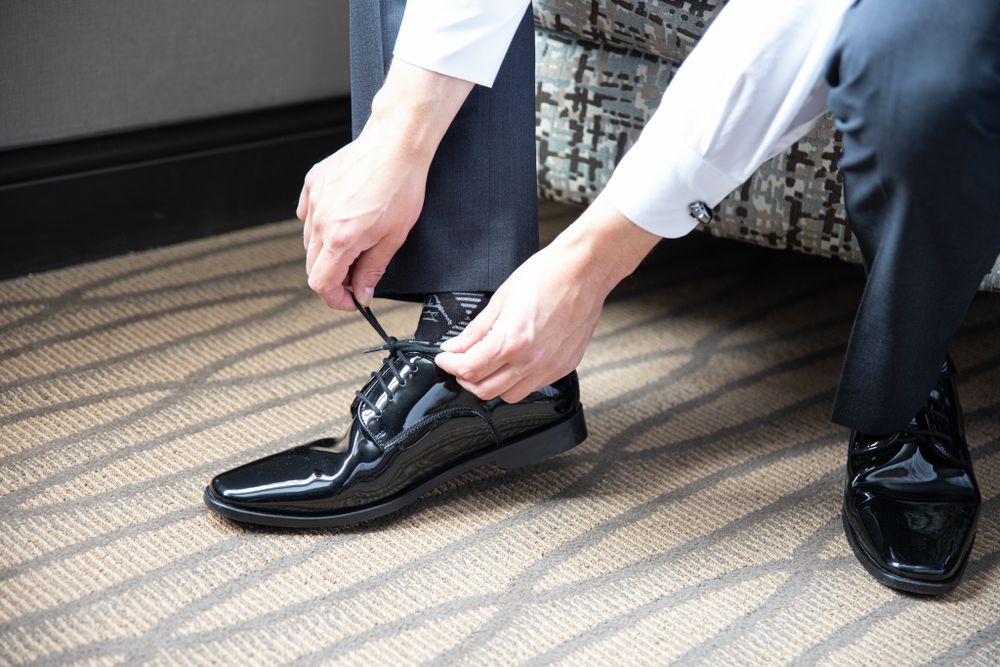 Close-up of someone tying shiny black formal dress shoes on carpeted floor.