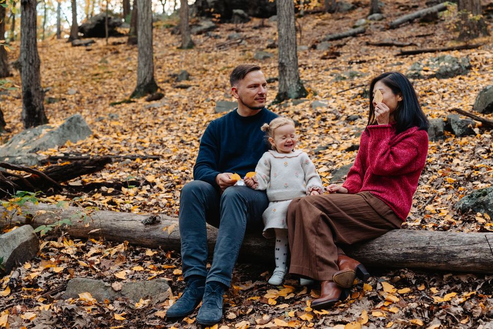 A family sits together on fallen logs in an autumn forest setting at Menotomy Rocks Park in Arlington, MA.