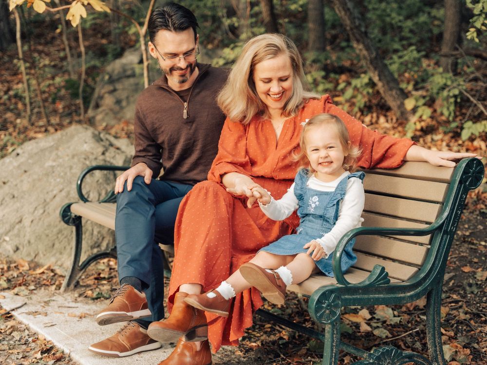 Family enjoying a playful autumn photoshoot outdoors on a wooden bench surrounded by fallen leaves at Menotomy Rocks Park in Arlington, MA.