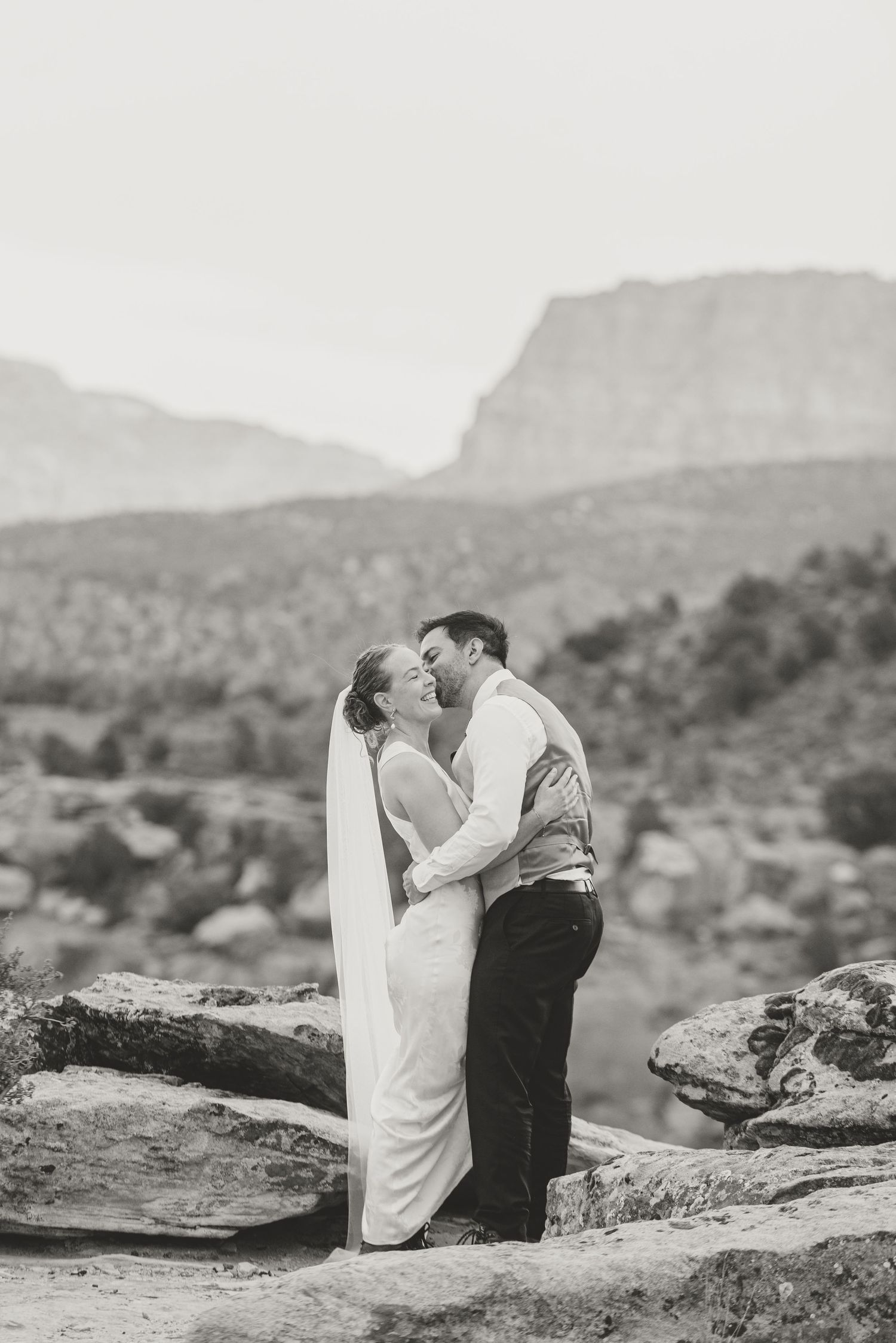 A couple stands in tall mountain grass surrounded by evergreen trees.