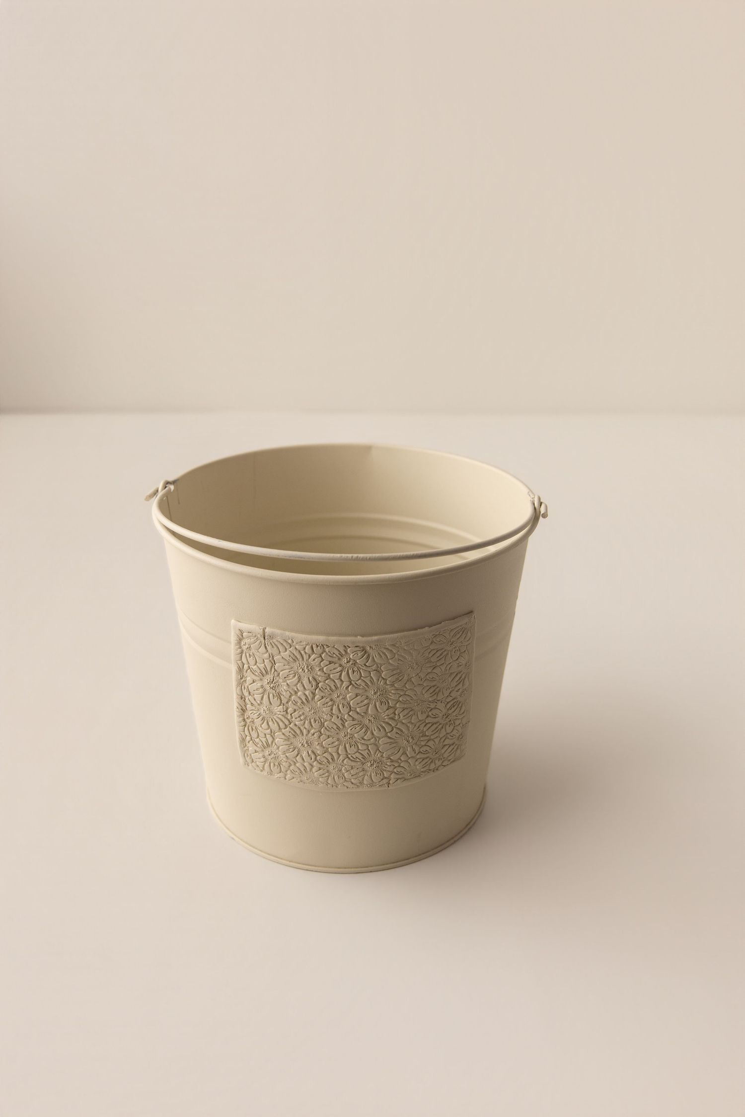 Cream metal bucket with decorative floral pattern, minimalist design, perfect for storage or floral arrangements.