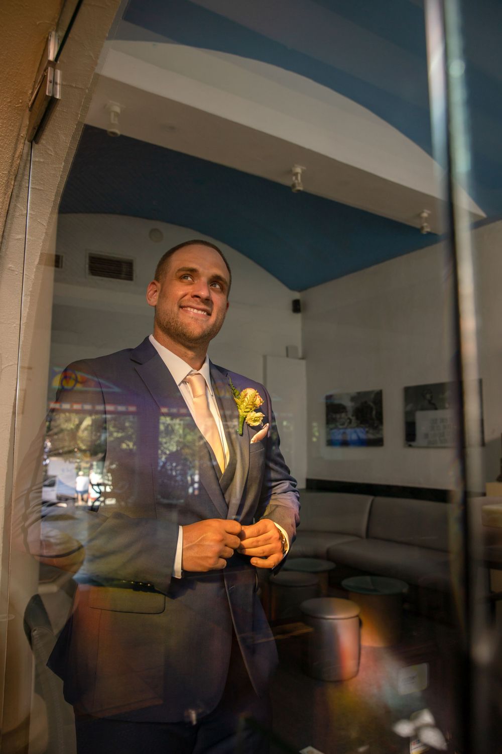 Person in a navy suit with yellow boutonniere adjusting their jacket in a room.