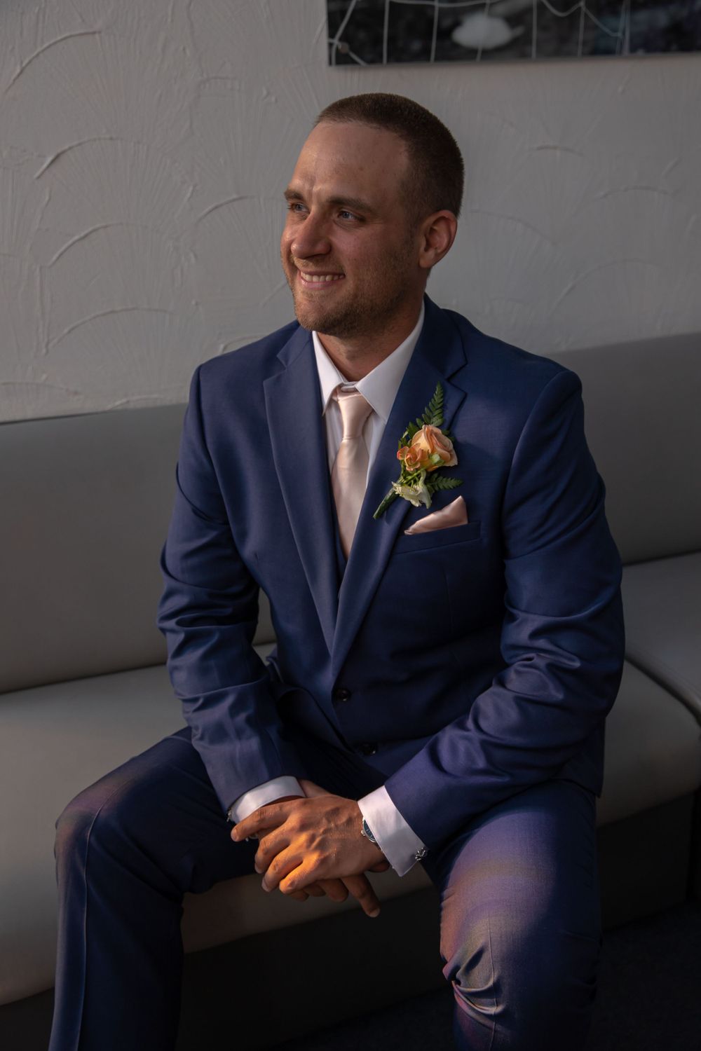 Person wearing a navy suit with peach tie and floral boutonniere sitting on grey couch.