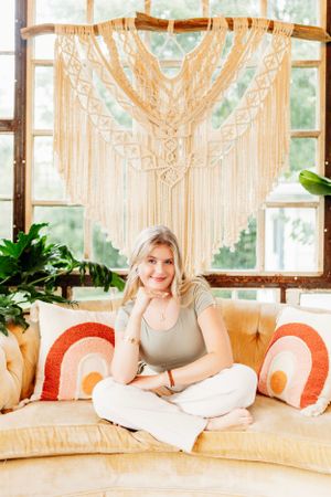Blonde woman in sage top sitting cross-legged on a cream couch beneath a macrame wall hanging.