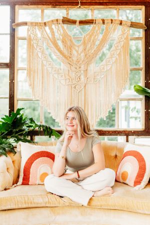 Blonde woman sitting cross-legged on a boho couch beneath a large macrame wall hanging indoors.