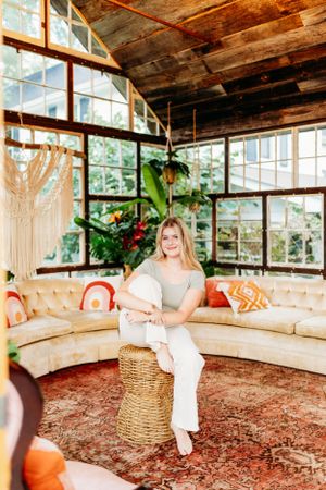 Smiling blonde woman seated on a wicker stool in a bohemian sunroom with lush tropical plants.