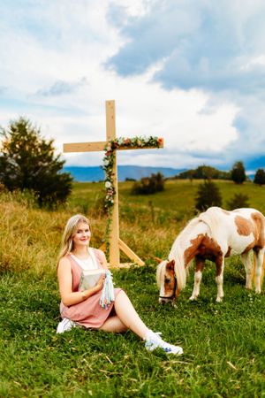 Blonde teen in pink dress sitting on grass reading a book beside a horse and wooden cross.
