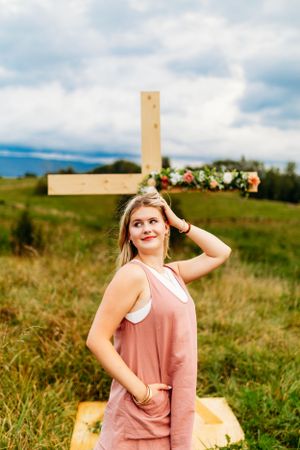 Smiling blonde girl in pink romper posing in front of a floral wooden cross in a green field.
