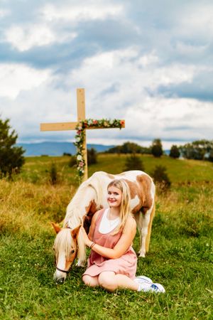 Blonde girl in pink dress sitting on grass beside a paint horse near a floral wooden cross.