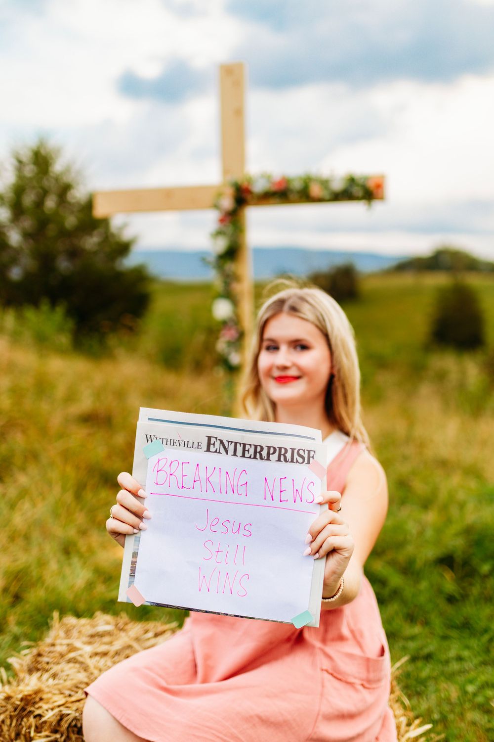 Young woman holding newspaper reading Breaking News Jesus Still Wins near a floral wooden cross.