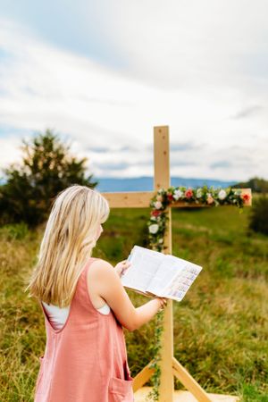 Blonde girl in pink overalls reading Bible beside a flower-adorned wooden cross in field.