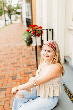 Blonde teen girl in cream top sitting on steps of brick sidewalk storefront.