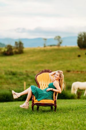 Blonde woman in green dress relaxing in vintage gold armchair on green farm pasture.