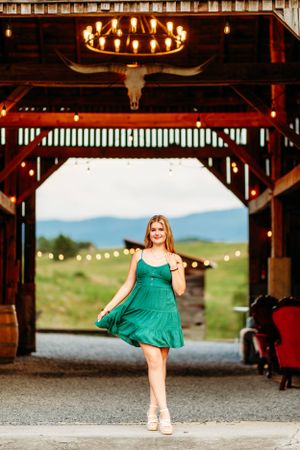 Young woman in green dress posing in rustic barn doorway with mountain views and string lights.