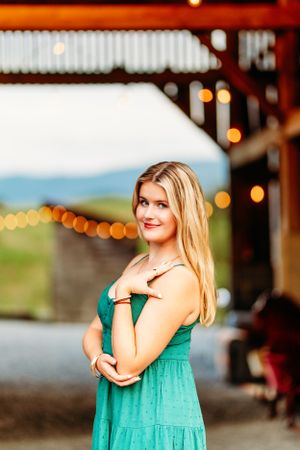 Smiling blonde teen in teal dress posing with arms crossed under string lights at barn.