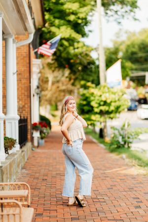 Teen girl in cream top and jeans walking on brick sidewalk near American flag.
