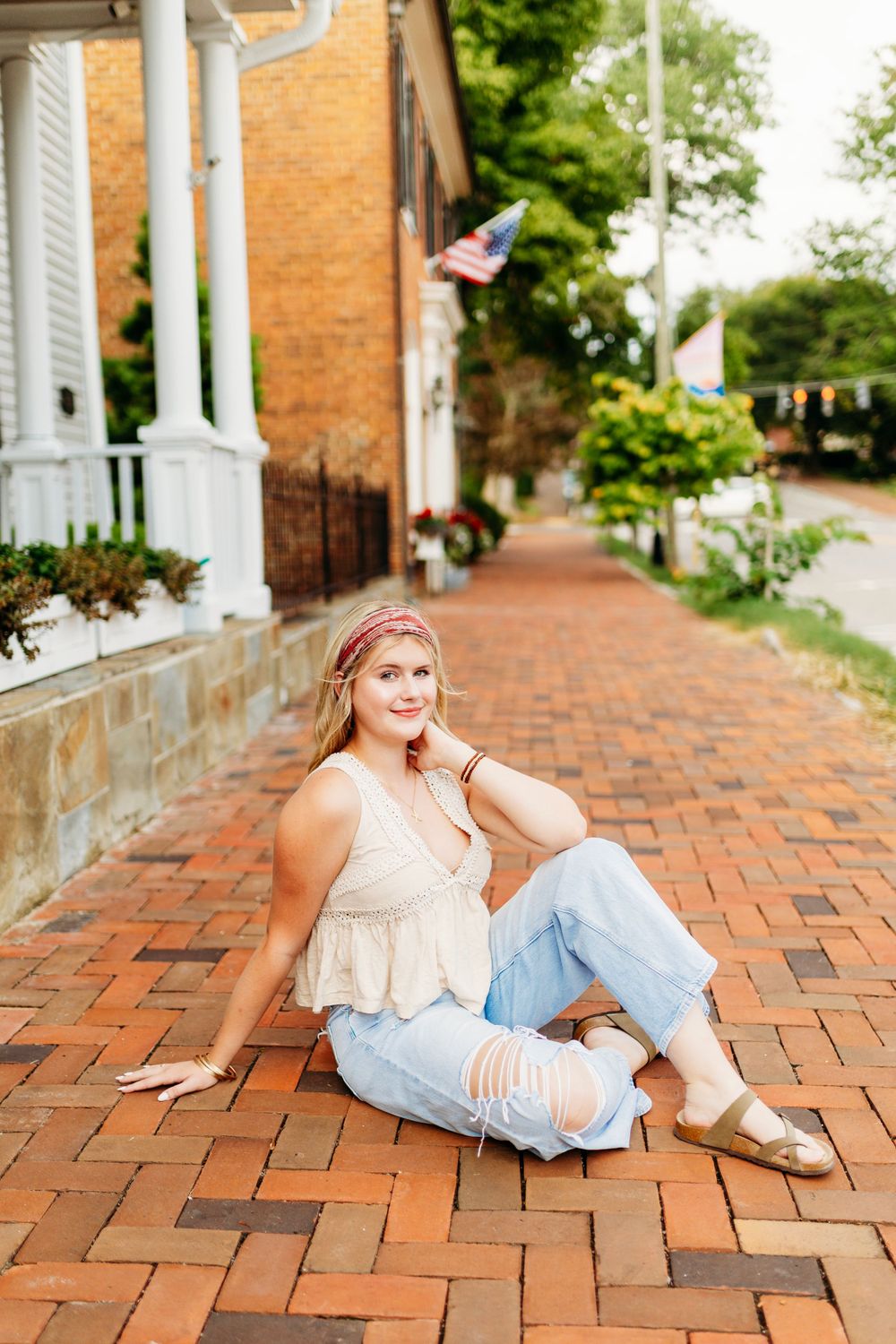 Smiling blonde teen in ripped jeans sitting on brick sidewalk near historic buildings.