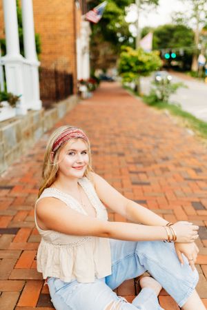 Smiling teen girl in ripped jeans sitting on brick sidewalk with American flag.
