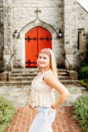 Blonde teen in boho top and jeans posing in front of a stone church with a red arched door.