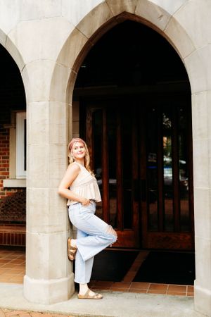 Young woman in light jeans and cream top leaning against stone arch doorway.