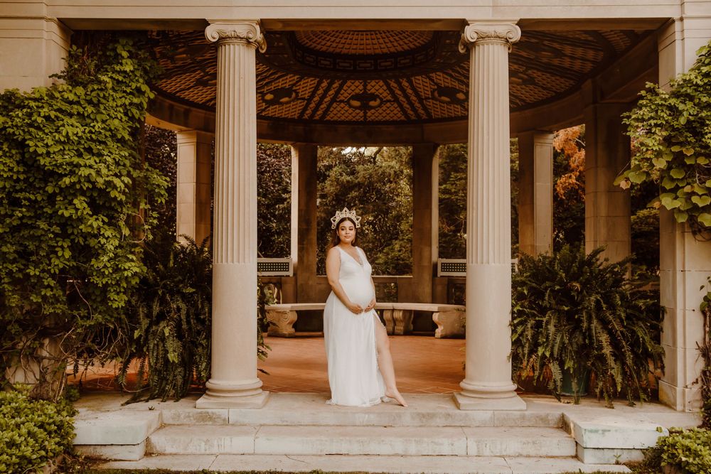 A white flowing dress stands elegantly in a classical garden gazebo with ornate columns.