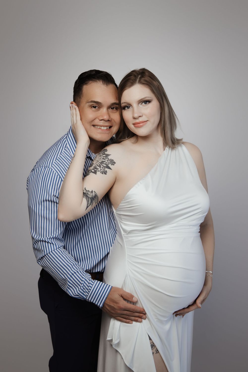 A couple poses for a maternity photo, woman in white dress with tattoos, man in blue striped shirt, gray background.
