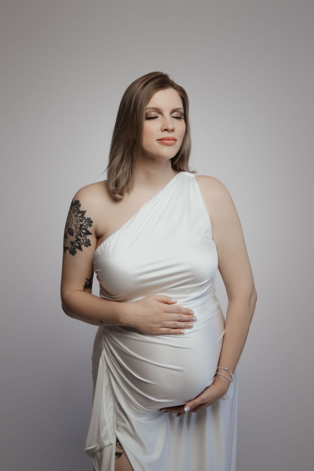 Pregnant woman in white one-shoulder dress with floral tattoo cradling baby bump in studio portrait.