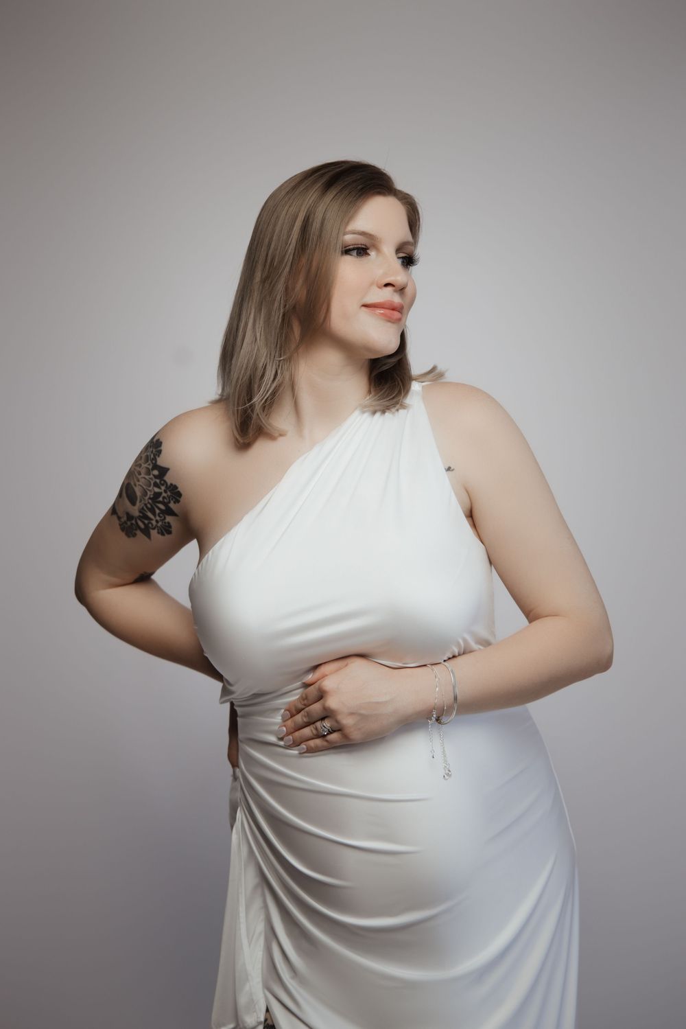 Woman in white one-shoulder dress with floral tattoo posing confidently against a light gray background.