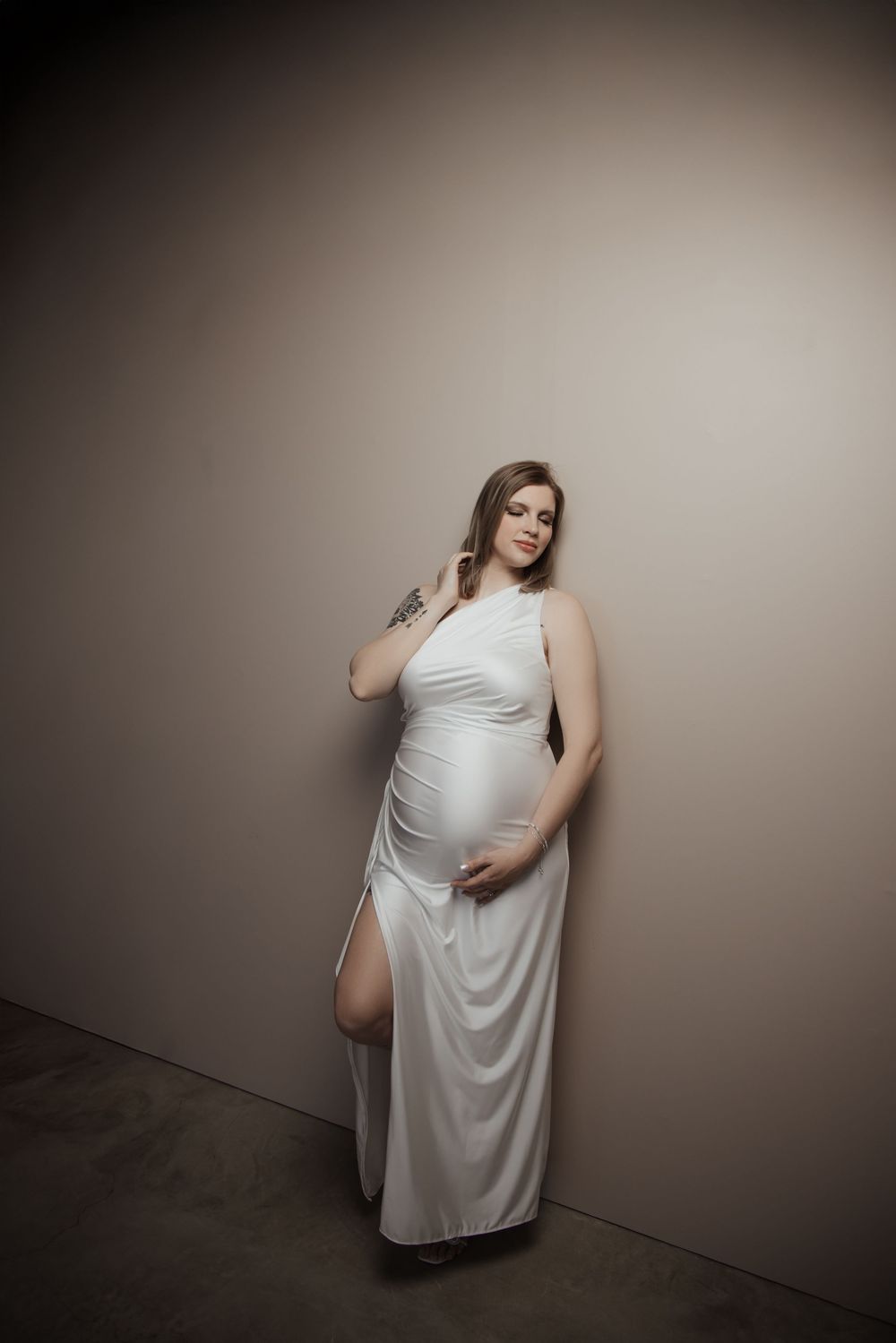 Pregnant woman in white halter maxi dress with slit poses gracefully against neutral wall, showcasing baby bump.