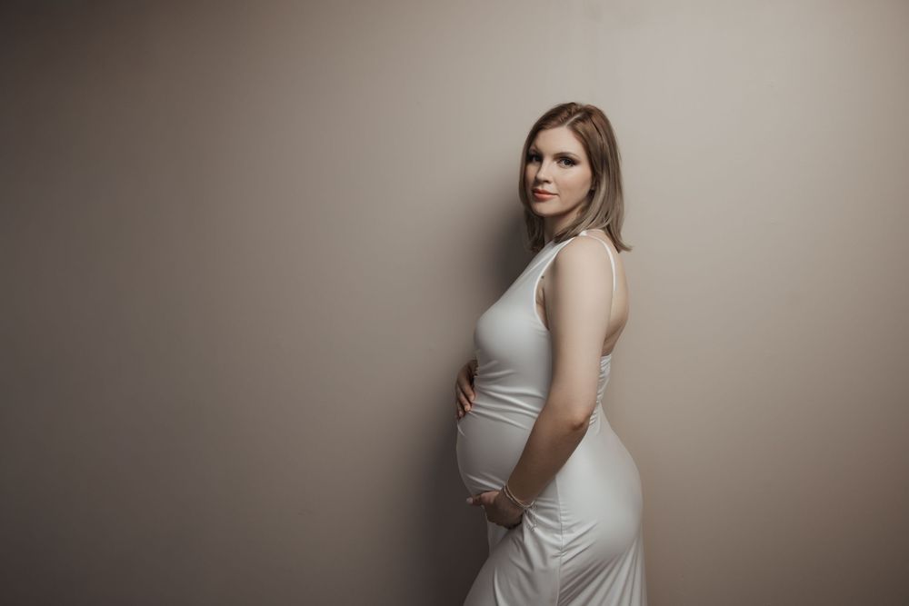 Pregnant woman in white dress cradling baby bump against beige wall in elegant maternity portrait photo.