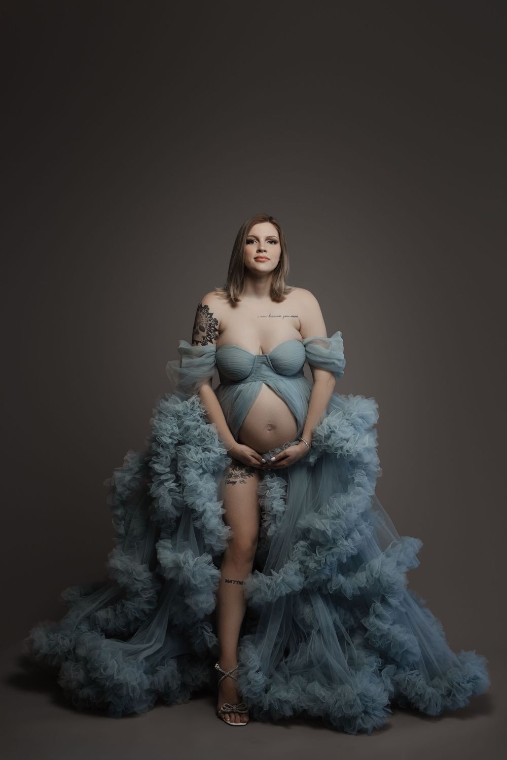Pregnant woman in a flowing blue tulle maternity gown cradling her baby bump in a studio portrait.