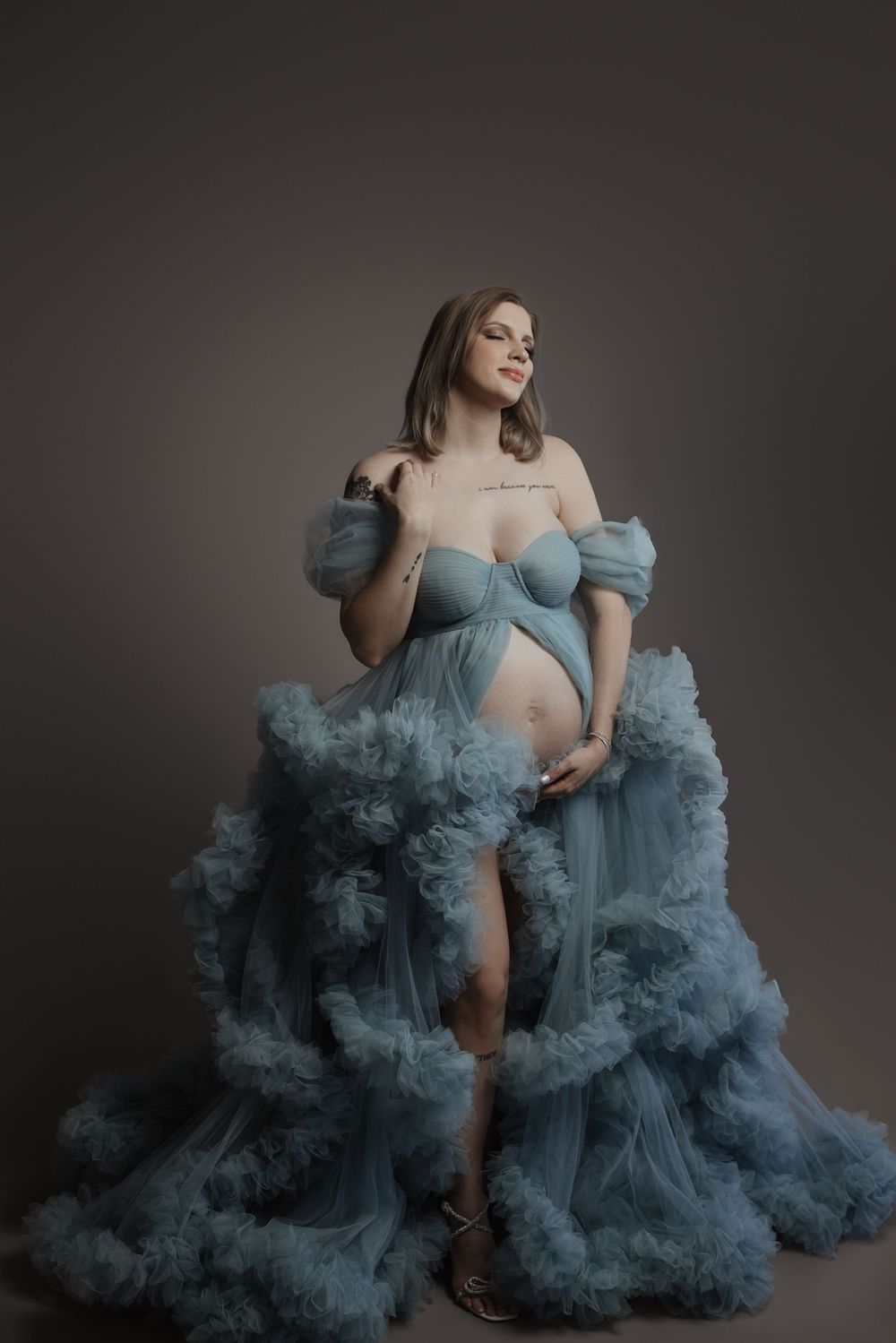 Pregnant woman in a flowing dusty blue off-shoulder tulle maternity gown poses gracefully in a studio setting.