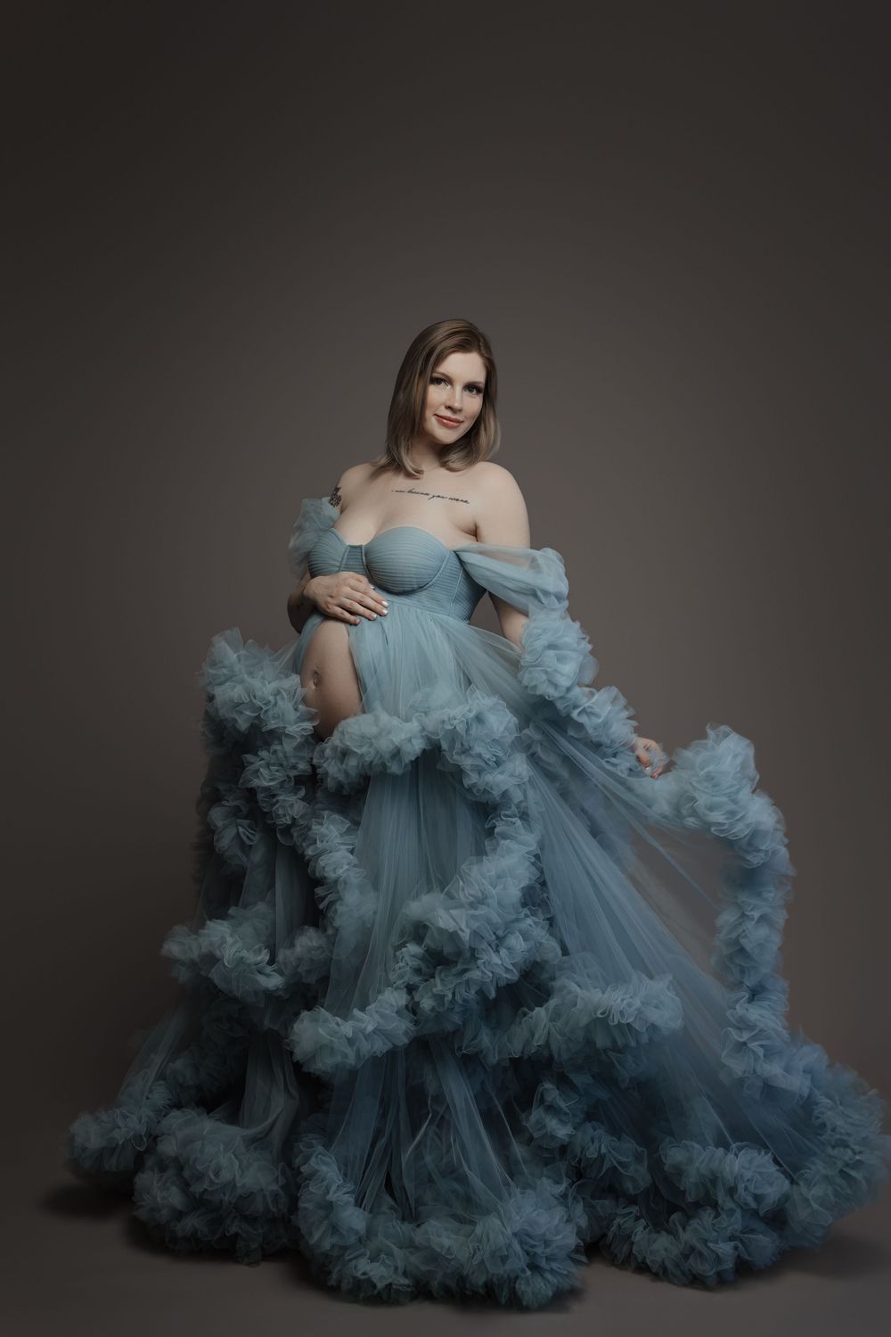 Pregnant woman in voluminous blue tulle off-shoulder gown posing gracefully in studio.