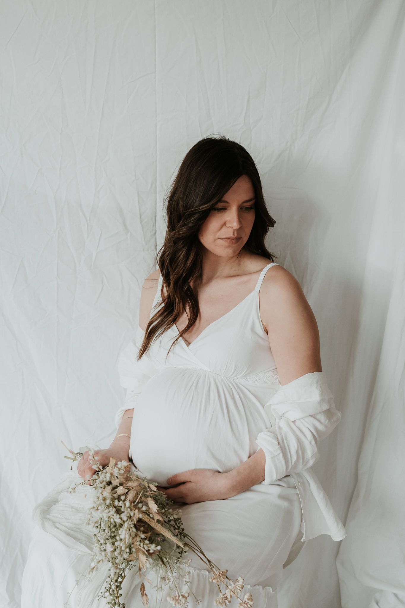 Artistic maternity portrait in flowing white dress with dried flowers.