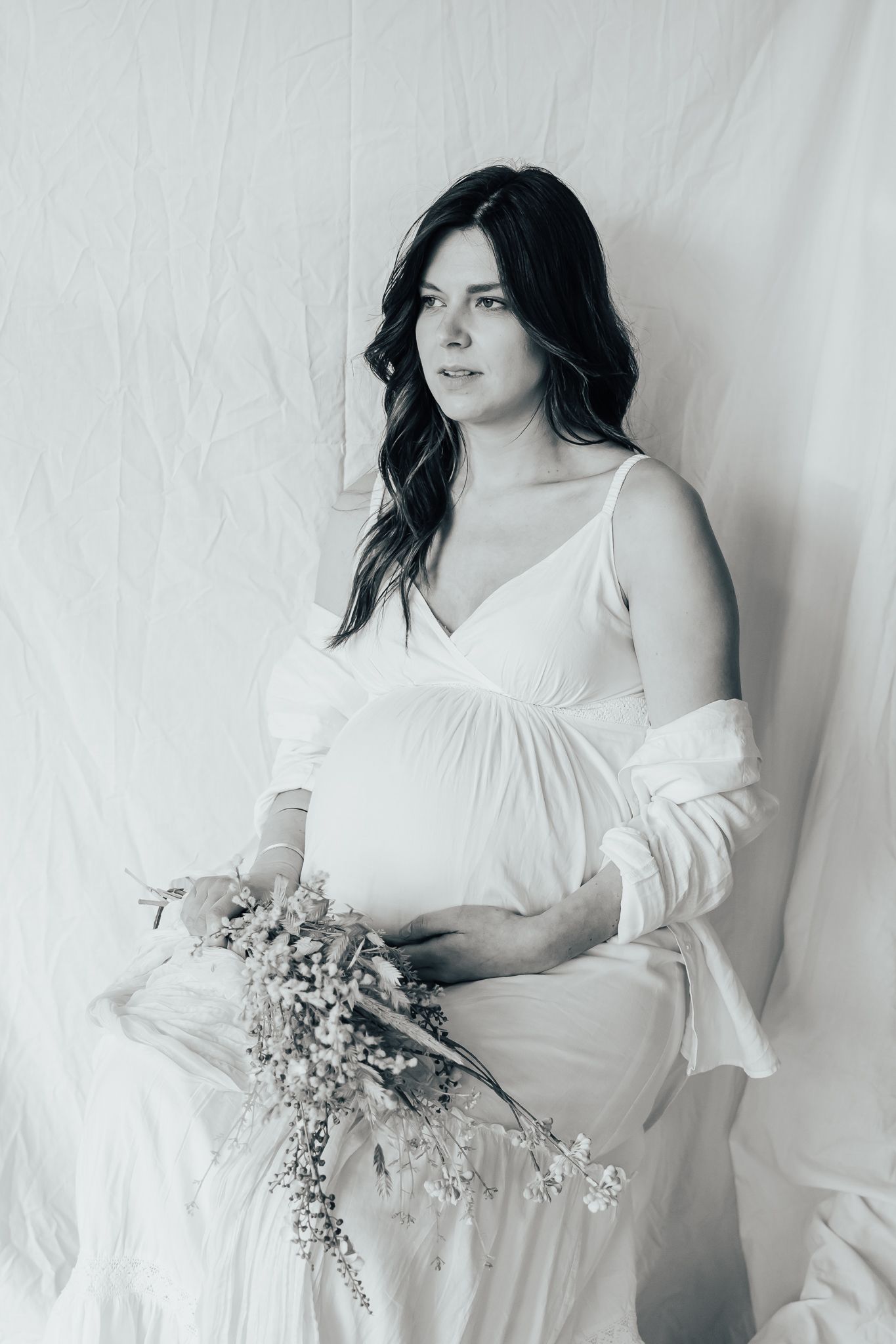 Maternity portrait in white dress with dried floral bouquet against light background.