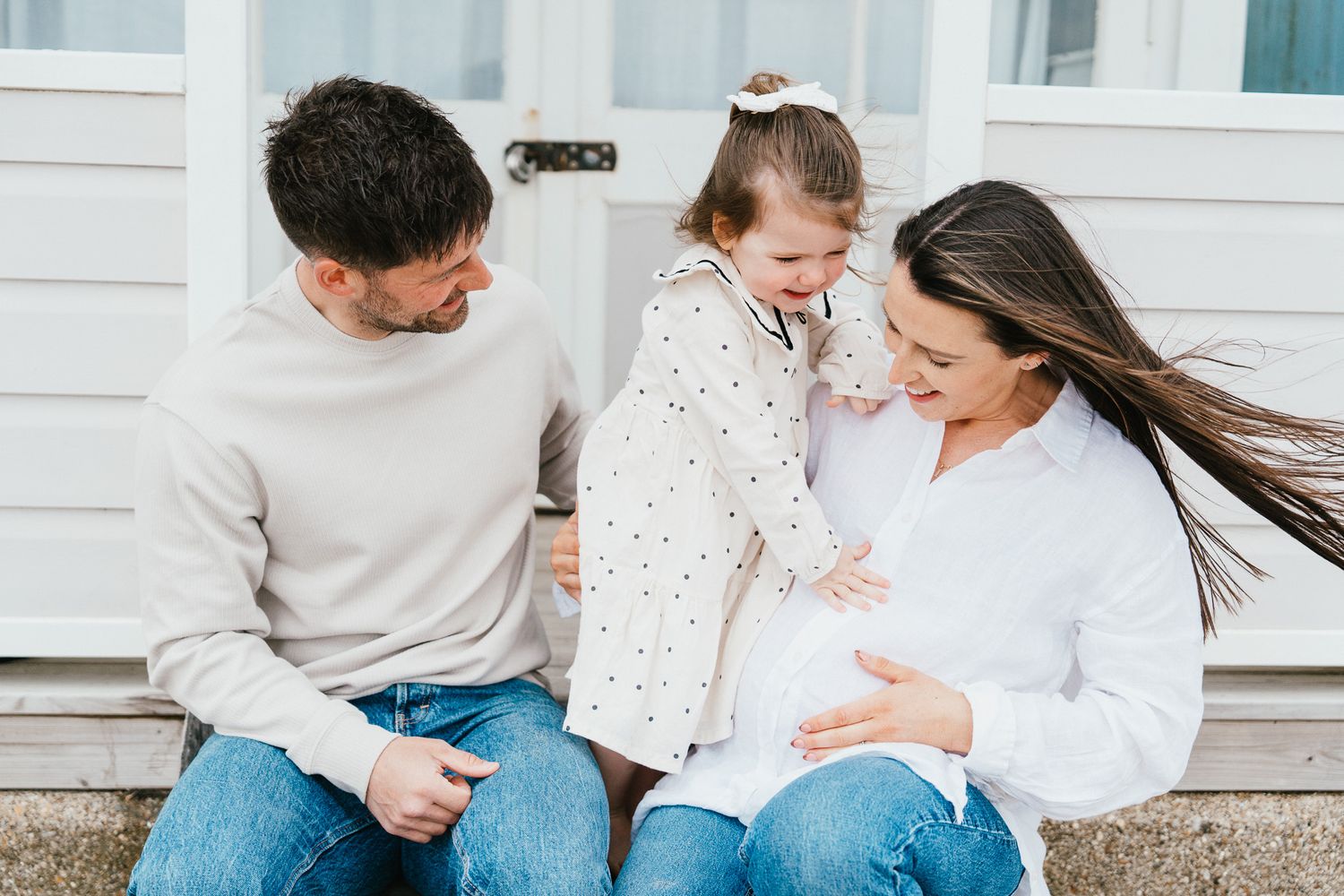 Happy family with young child laughing together outside white building on windy day.