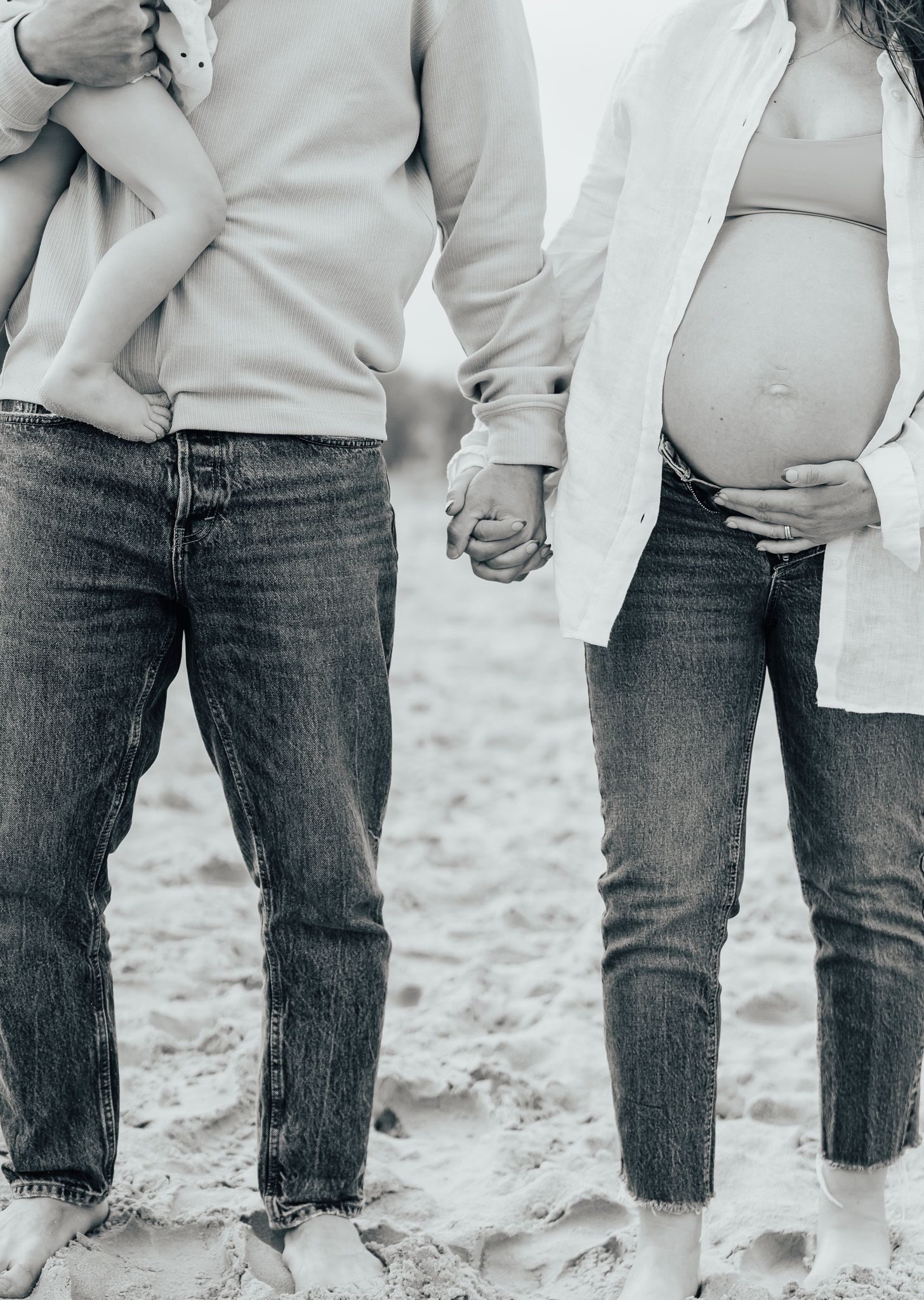 Black and white photo of couple holding hands on sandy beach showing baby bump.