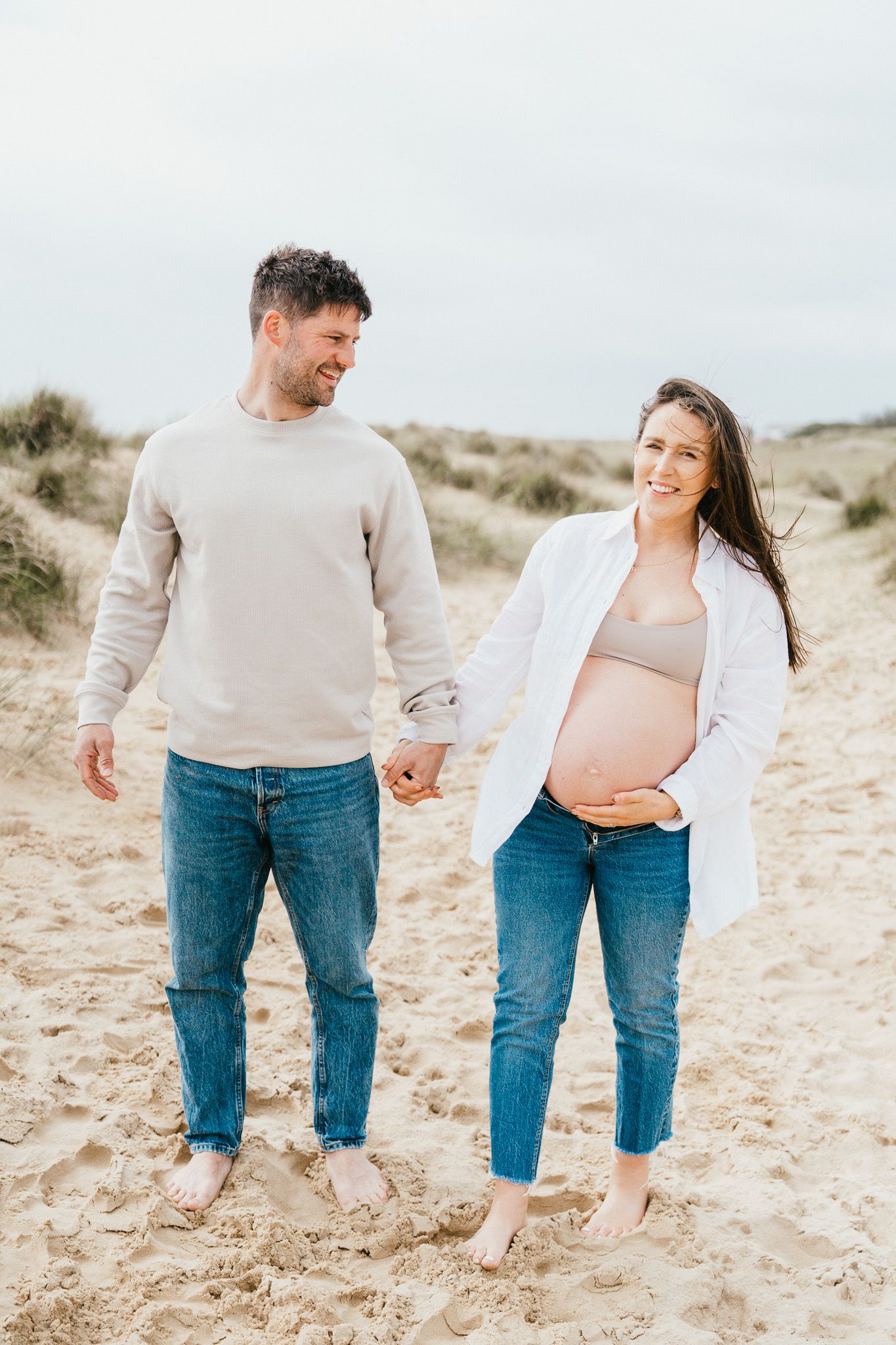 Couple walking barefoot on sandy dunes during maternity photoshoot.