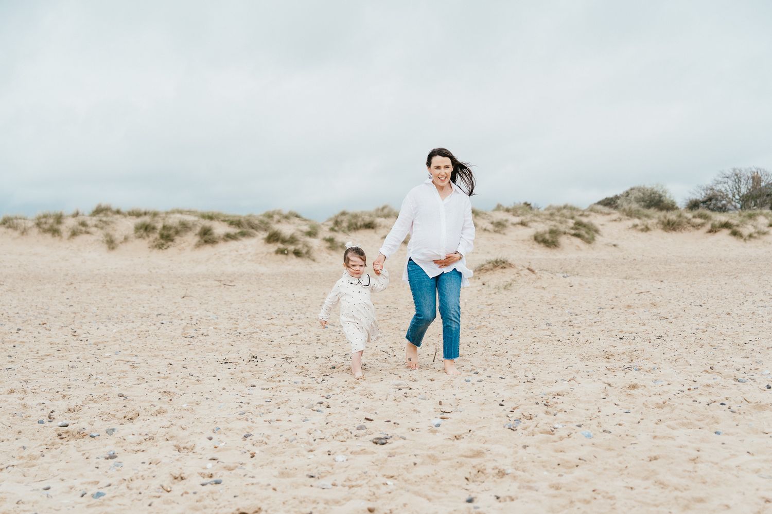 Mother and child running together on empty beach during overcast day.
