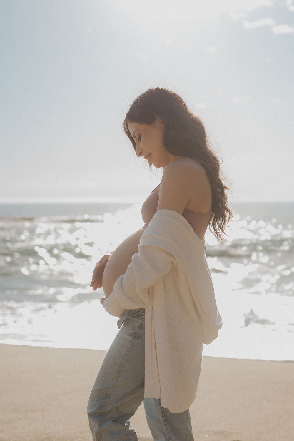 A woman in white stands thoughtfully by the ocean shoreline.