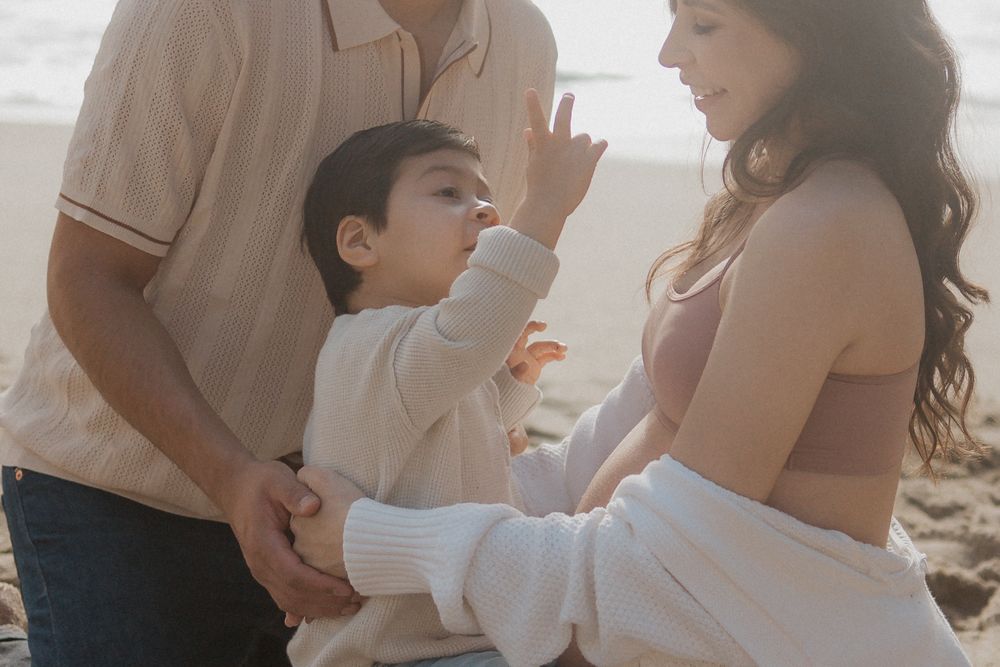 Intimate family moment on beach with toddler reaching up during maternity shoot.