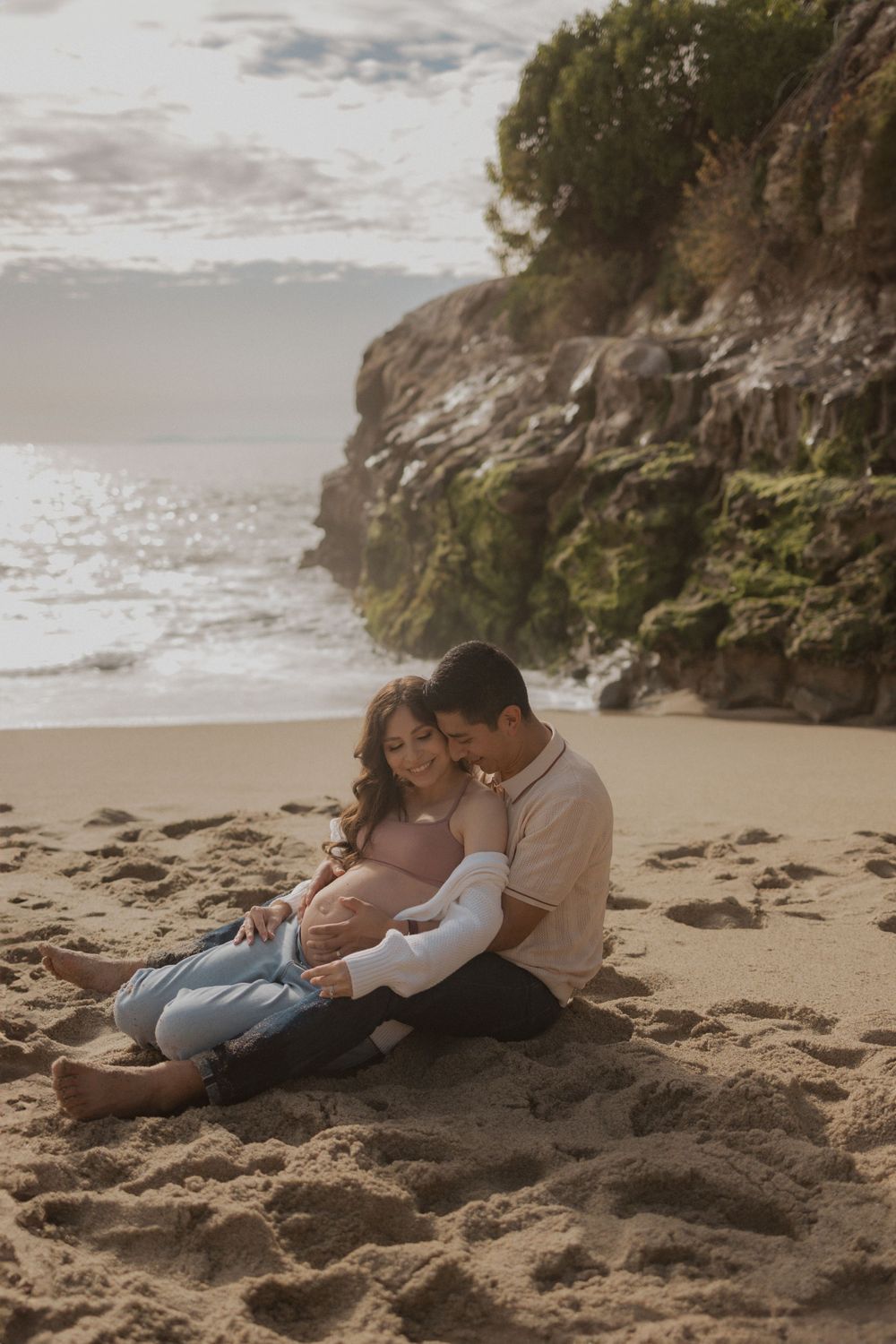 Romantic maternity photo of couple embracing on scenic beach with cliffs.