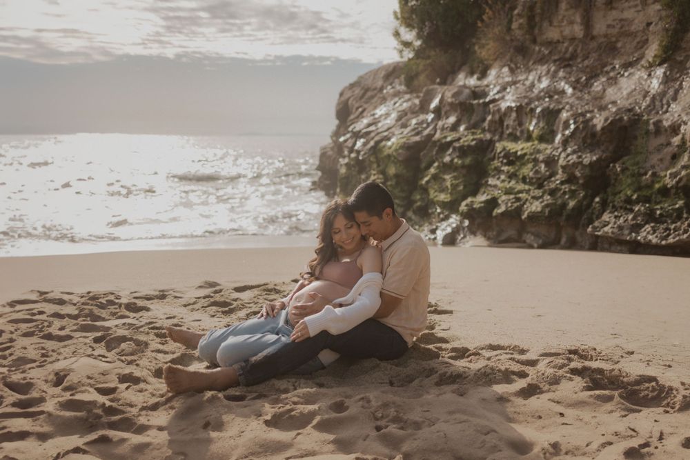 A couple sits intimately on a sandy beach near rocky cliffs at sunset.