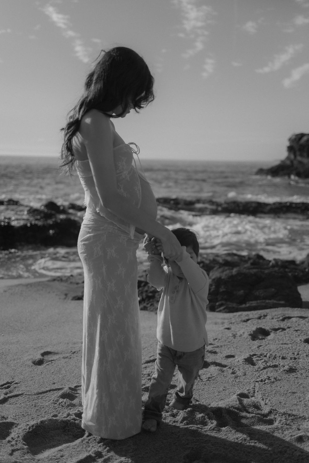 A pregnant woman in a long dress stands with a small child on a rocky beach at sunset.