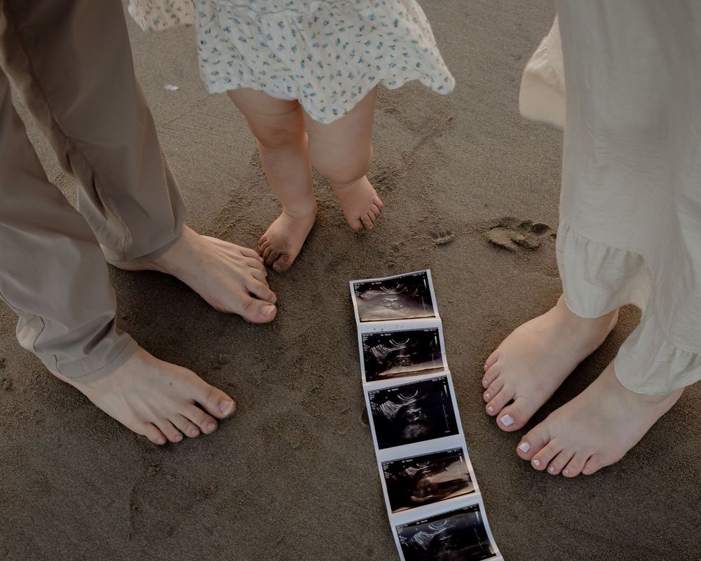 Barefoot family standing on sandy beach around ultrasound photos, pregnancy announcement with child and parents feet.
