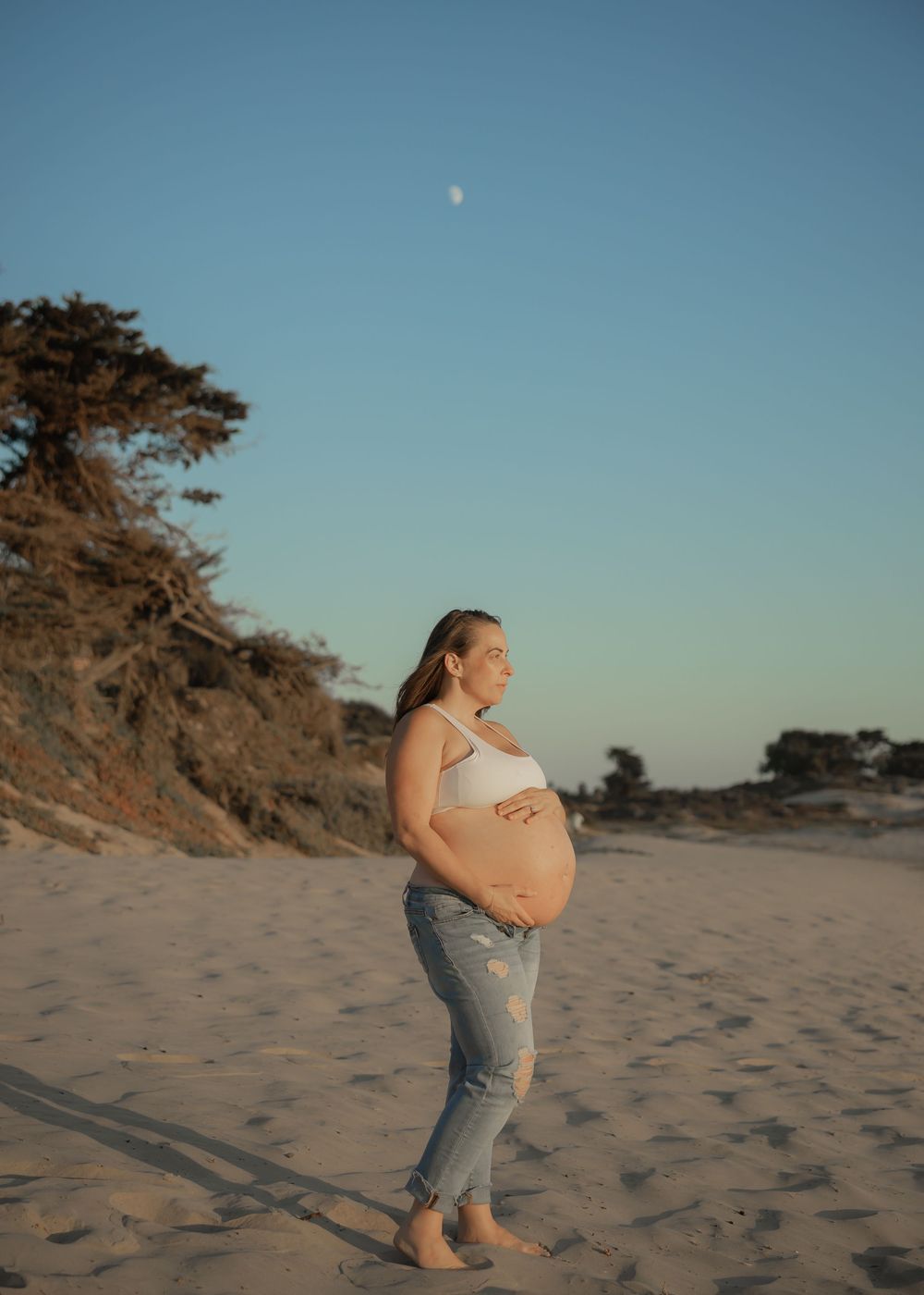 Pregnant woman in white crop top and jeans cradling baby bump on sandy beach at golden hour with moon visible.