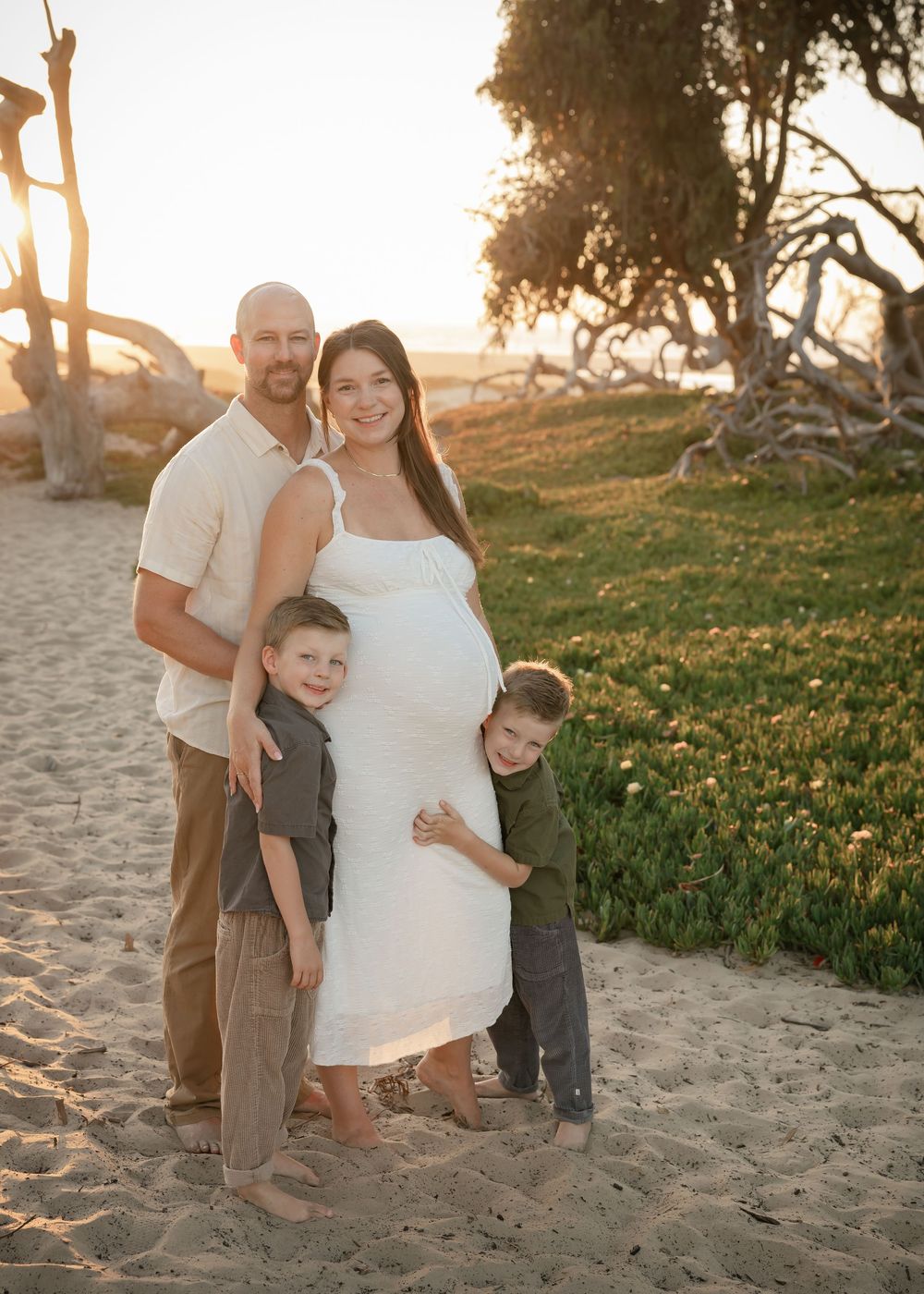 Pregnant woman in white dress poses with husband and two young sons on sandy beach at golden hour.